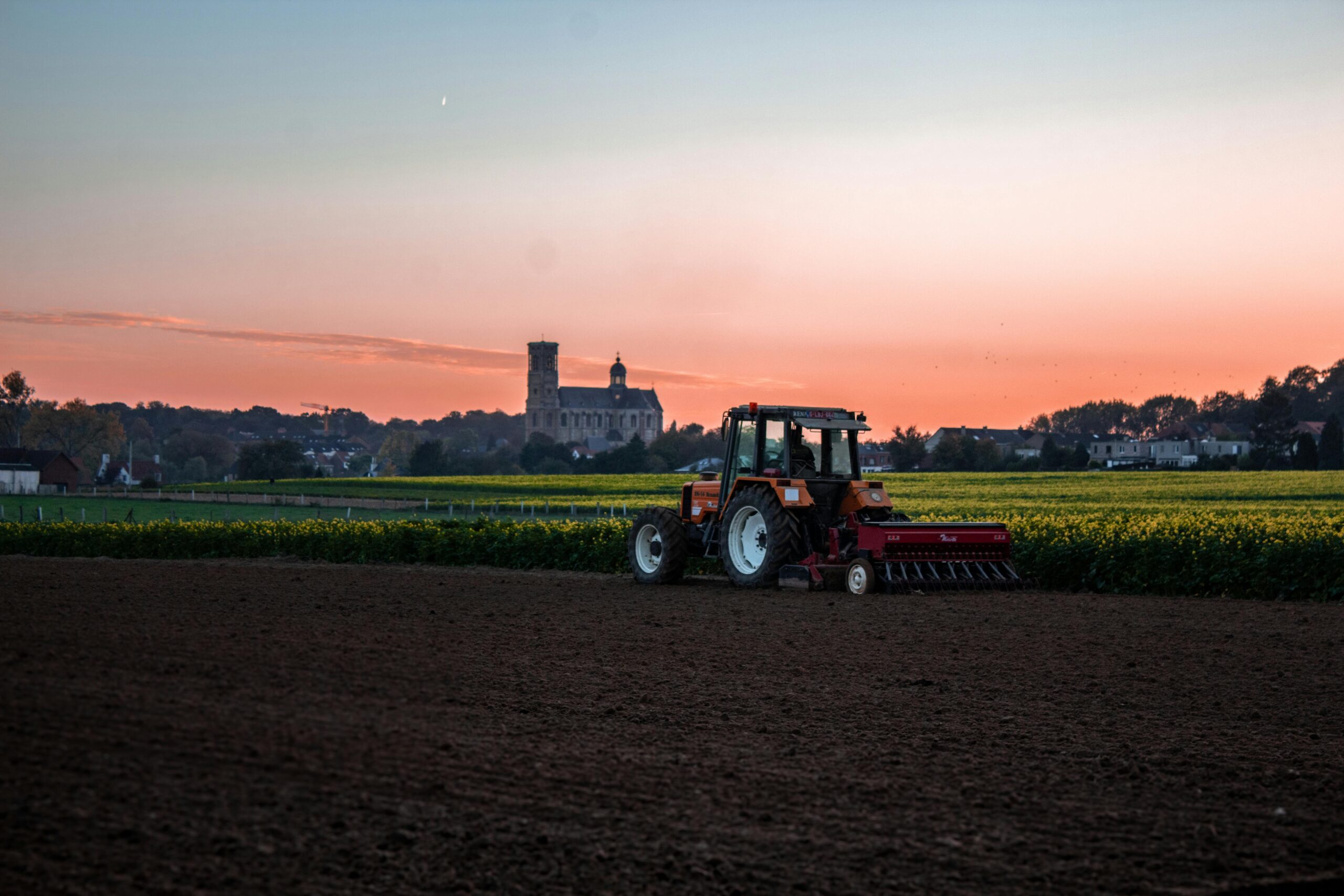 A red tractor plows a field under a vibrant sunset near a countryside landmark.