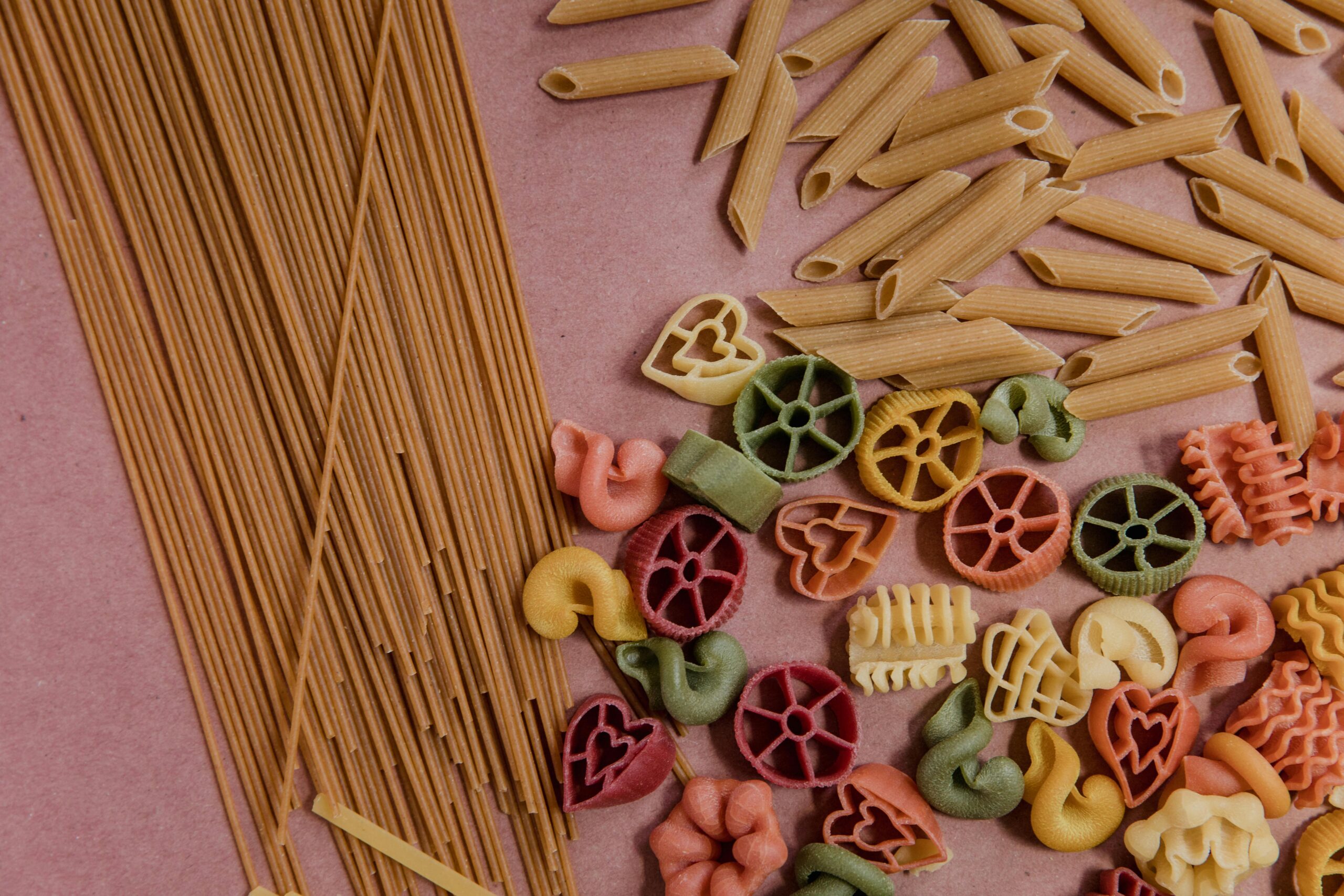 Vibrant assortment of uncooked pasta in various shapes and colors, displayed on a pink surface